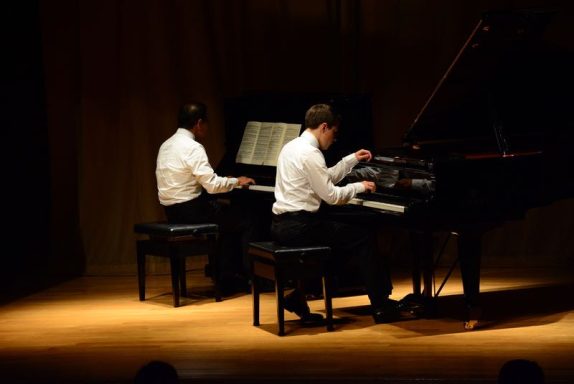 Zwei Pianisten spielen synchron auf einem Klavier auf einer Bühne.
